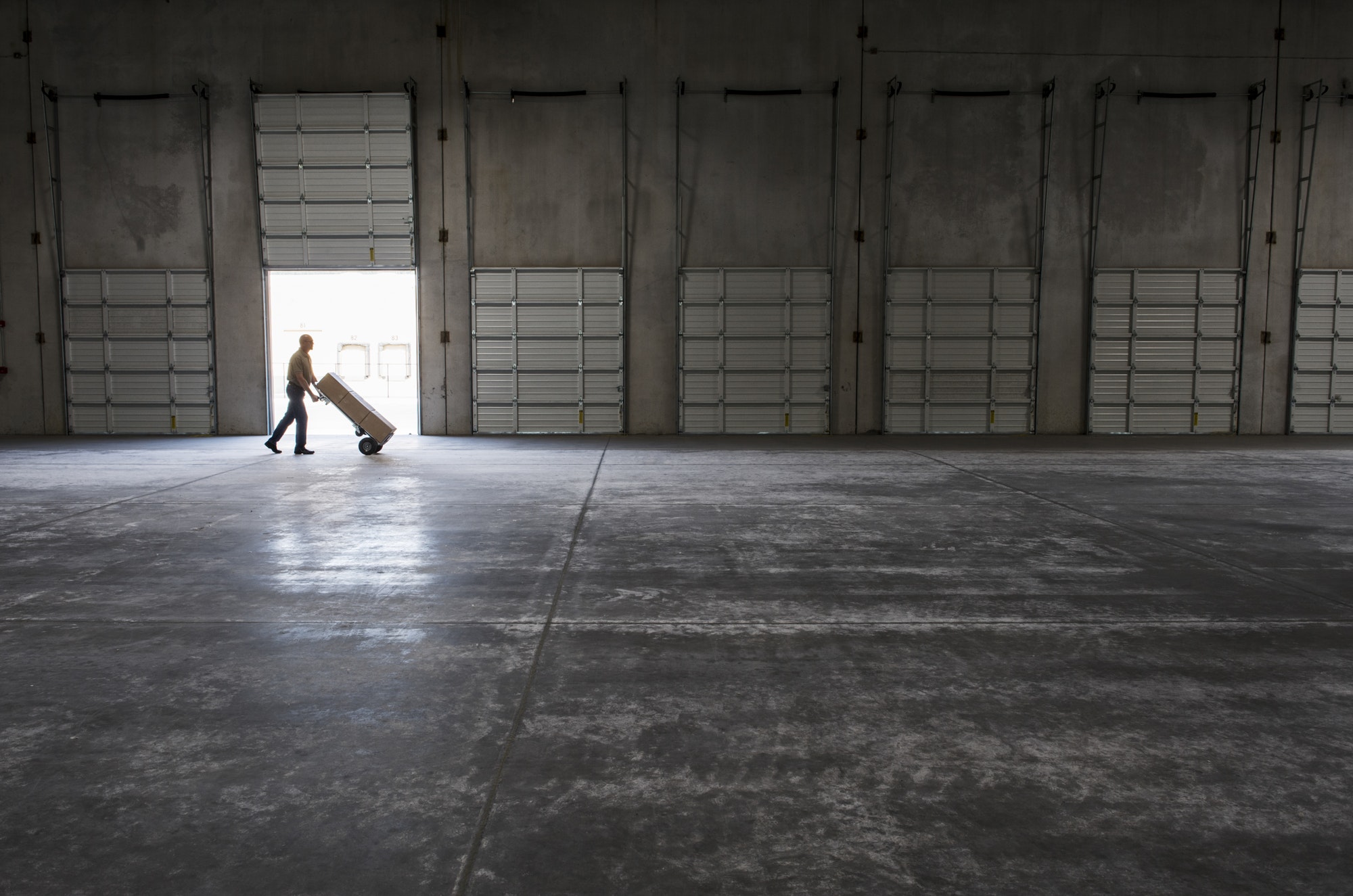 Man walking with loaded hand truck past loading dock doors in new empty warehouse.